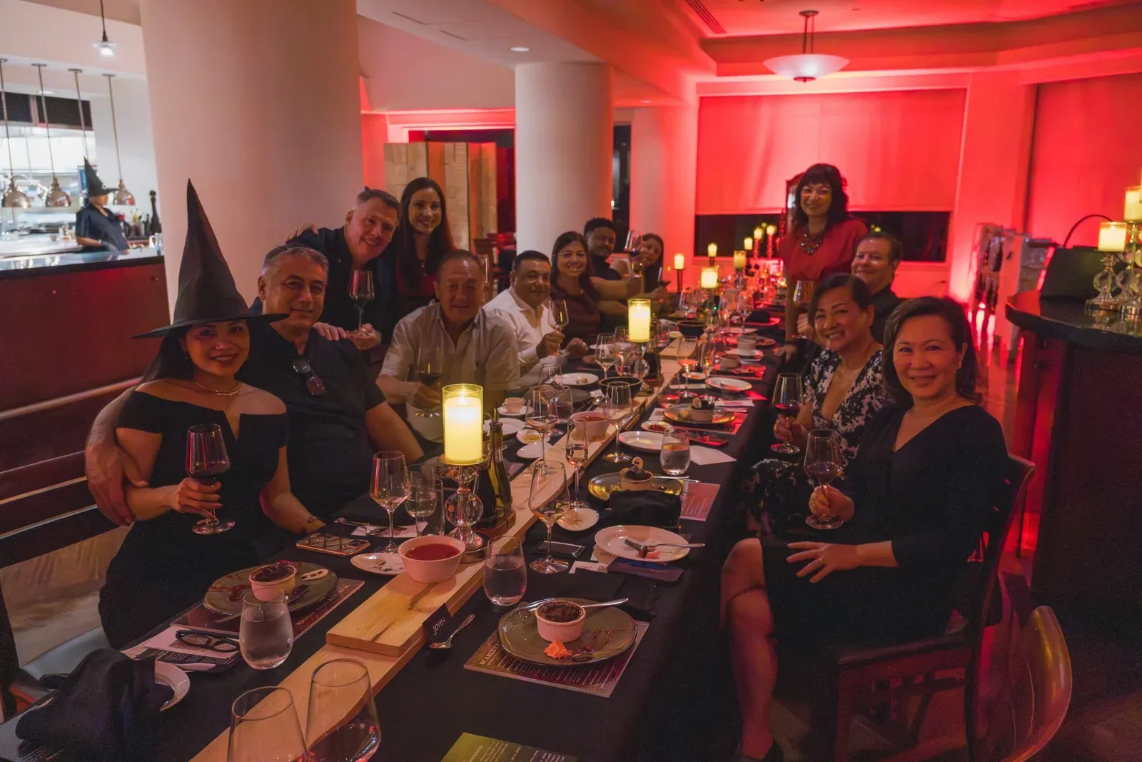 Guests, one in a witch hat, at a long dinner table lit red for Halloween
